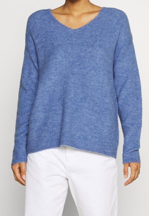 Personne portant un pull bleu à manches longues, ample et doux au toucher, avec un col en V et un pantalon blanc, mains détendues le long du corps.