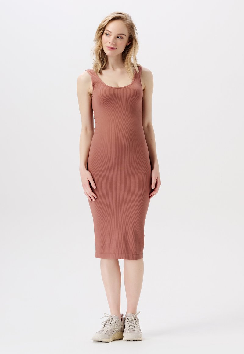 Kvinna som står klädd i en ärmlös, rippad, knälång klänning i dammrosafärg och beige sneakers, med blicken något vänd åt sidan mot en vit bakgrund.