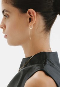 Aretes geométricos en tono dorado con una caída de cadena, combinados con una blusa de cuero negro elegante. Los aretes presentan un acento redondo y una barra alargada.