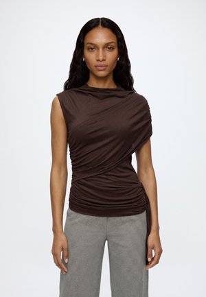 Femme aux longs cheveux ondulés, portant un haut froncé sans manches marron foncé et un pantalon à carreaux gris clair, se tenant devant un fond blanc.
