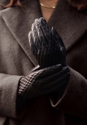 Schwarze, gesteppte Lederhandschuhe mit strukturiertem Oberflächen, die eine enganliegende Form und verlängerte Finger aufweisen, gehalten von einer Person in einem dunkelgrauen Mantel.