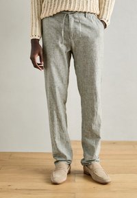 Light gray linen pants with a loose fit, drawstring waist, and side pockets, paired with beige suede loafers. Wood floor background.