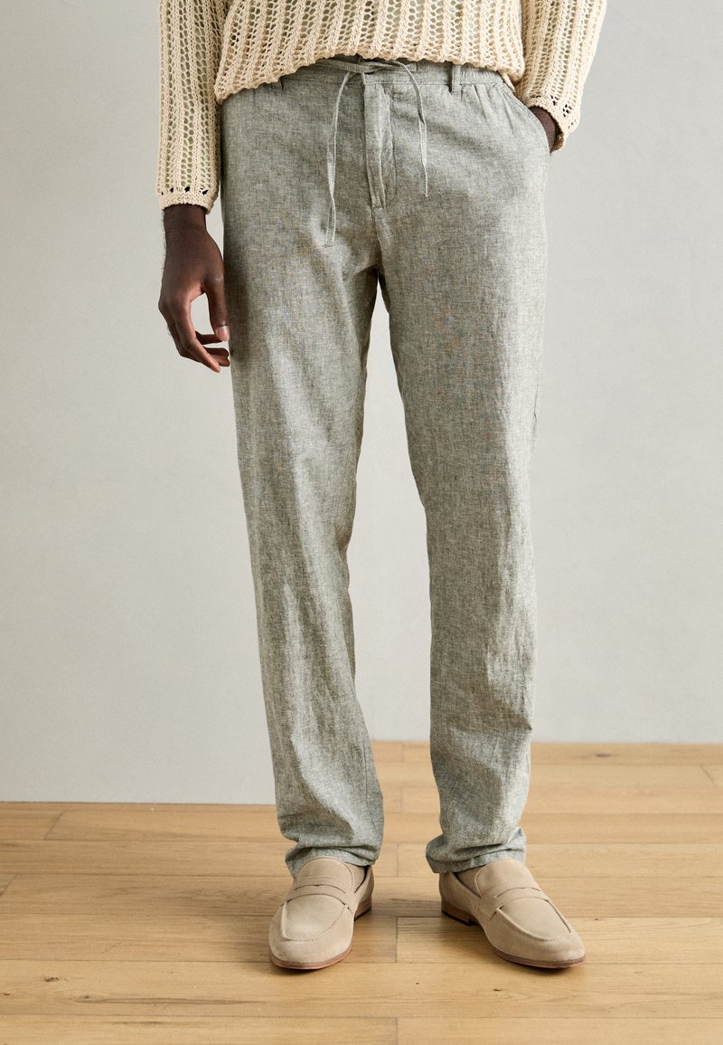 Light gray linen pants with a loose fit, drawstring waist, and side pockets, paired with beige suede loafers. Wood floor background.