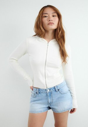 Junge Frau mit langen braunen Haaren, die einen weißen Kapuzenpullover mit Reißverschluss und hellblaue Jeansshorts trägt, steht vor einem einfarbigen weißen Hintergrund.