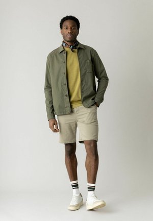 Mand stående iført grøn skjortejakke, gul sweater, beige shorts, mønstret tørklæde om halsen, hvide sneakers og stripede strømper, neutral baggrund.
