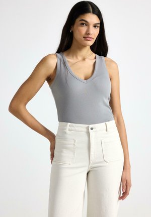 Kvinde med langt mørkt hår iført grå ærmeløs V-hals top og højtaljede off-white bukser, stående med den ene hånd på hoften.