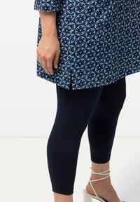 Tunique à motif floral bleu et blanc associée à un legging en marine ajusté, présentant une fente sur le côté et une texture de tissu légère. Sandales à talons beiges.