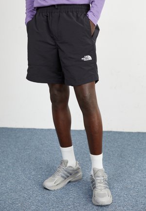 Personne portant un short noir The North Face, des chaussettes blanches et des baskets Adidas grises, debout sur un sol recouvert d'un tapis bleu contre un mur blanc.