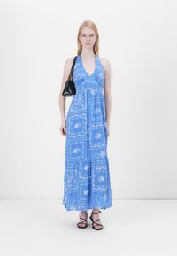 Blaues Maxikleid mit floralen Muster, Halterneck-Design und gestuftem Saum. Das Model hält eine kleine schwarze Tasche und trägt dazu schwarze Sandalen mit Riemchen.