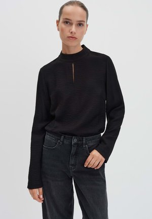 Donna che indossa un top nero a maniche lunghe con texture e una piccola fessura frontale, abbinato a jeans neri a vita alta, su uno sfondo chiaro e semplice.
