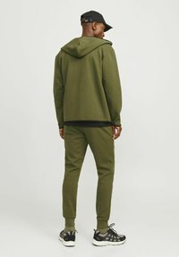 Olive green hoodie with a front pocket and matching tapered joggers. Black cap and athletic shoes complete the outfit. Soft, textured fabric.
