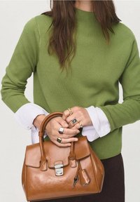 Sac à main en cuir marron de forme structurée, avec des accessoires en argent et un porte-clés détachable. Porté avec un pull vert et des poignets de chemise blancs.