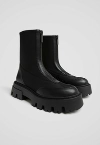 Bottines noires à semelle épaisse, fabriquées avec des matériaux lisses et texturés. Dotées de deux zips à l'avant et de détails robustes sur la semelle.