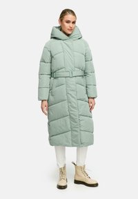 Puffig mintgrön lång kappa med huva, bälte i midjan och diamantmönstrad quiltning. Matchas med vita byxor och beige snörstövlar.