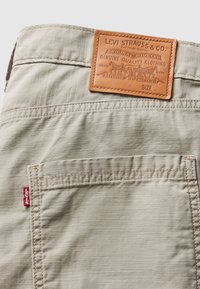 Bežene pamučne hlače s tamnosmešnim kožnim uzorkom embosiranim s "Levi Strauss & Co." i crvenom oznakom Levi's na džepu.