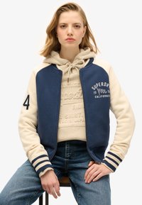 Marineblauwe en crèmekleurige varsityjas met geribde manchetten, voorzien van een ritssluiting aan de voorkant en geborduurde logo's, gedragen over een beige capuchontrui.