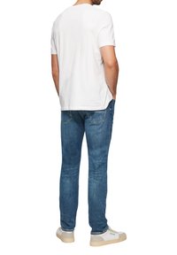 Witte kortekous t-shirt, blauwe spijkerbroek en beige sneakers, gezien vanaf de achterkant. De spijkerbroek heeft een klassieke pasvorm met zakken.