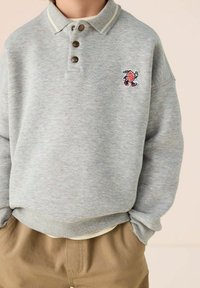 Grauer Pullover mit Kragen, drei braunen Knöpfen und einem bestickten Charakter. Besitzt einen strukturierten Stoff mit subtilen, mehrfarbigen Sprenkeln.