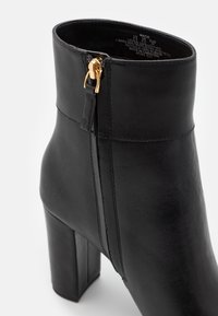 Black leather ankle boot with a chunky high heel, featuring a side zipper with gold hardware and a smooth texture.