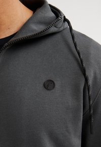 Sudadera gris con cordón, que presenta un parche de logo negro circular en la parte frontal. La tela tiene una textura suave, creando una apariencia relajada.