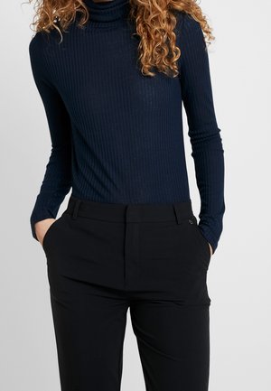 Femme portant un pull à col roulé côtelé bleu marine et un pantalon noir sur mesure, les mains dans les poches, cadrée des épaules vers le bas.
