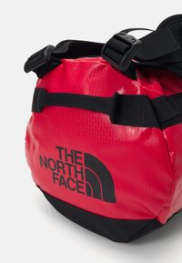 Red waterproof duffel bag with black accents and logo. Features adjustable straps, a textured exterior, and a rounded shape.