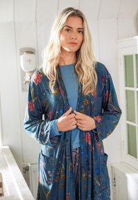 Blonde woman indoors wearing a long blue floral robe over a blue shirt, standing with hands clasping the robe front.