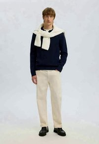 Tengerészkék kötött pulóver, krémszínű sállal megkötve a nyaknál, krémszínű nadrággal és fekete cipővel párosítva. Egyszerű háttér.