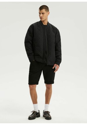 Młody mężczyzna stoi, ubrany w czarną pikowaną kurtkę bomber, czarne szorty, białe skarpetki i czarne sznurowane buty na neutralnym tle.