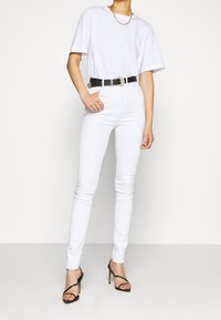 Jeans ajustados blancos de talle alto con una forma ceñida, combinados con una camiseta blanca holgada y un cinturón negro con una hebilla decorativa dorada.