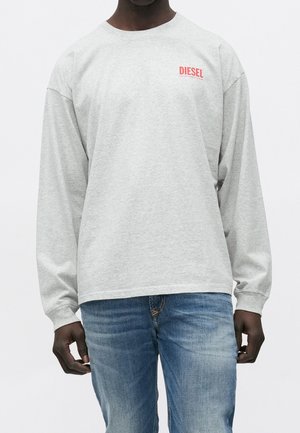 Hombre vistiendo una camiseta de manga larga Diesel gris claro con logo rojo en el pecho y vaqueros azules sobre fondo blanco liso.