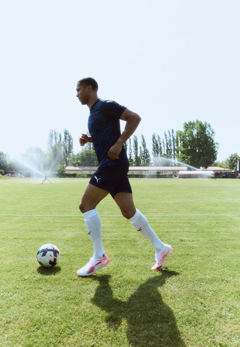 Joueur de football Puma dans un maillot à motifs bleu marine et des chaussettes blanches, courant sur de l'herbe avec un ballon noir et blanc, entouré de sprinklers.