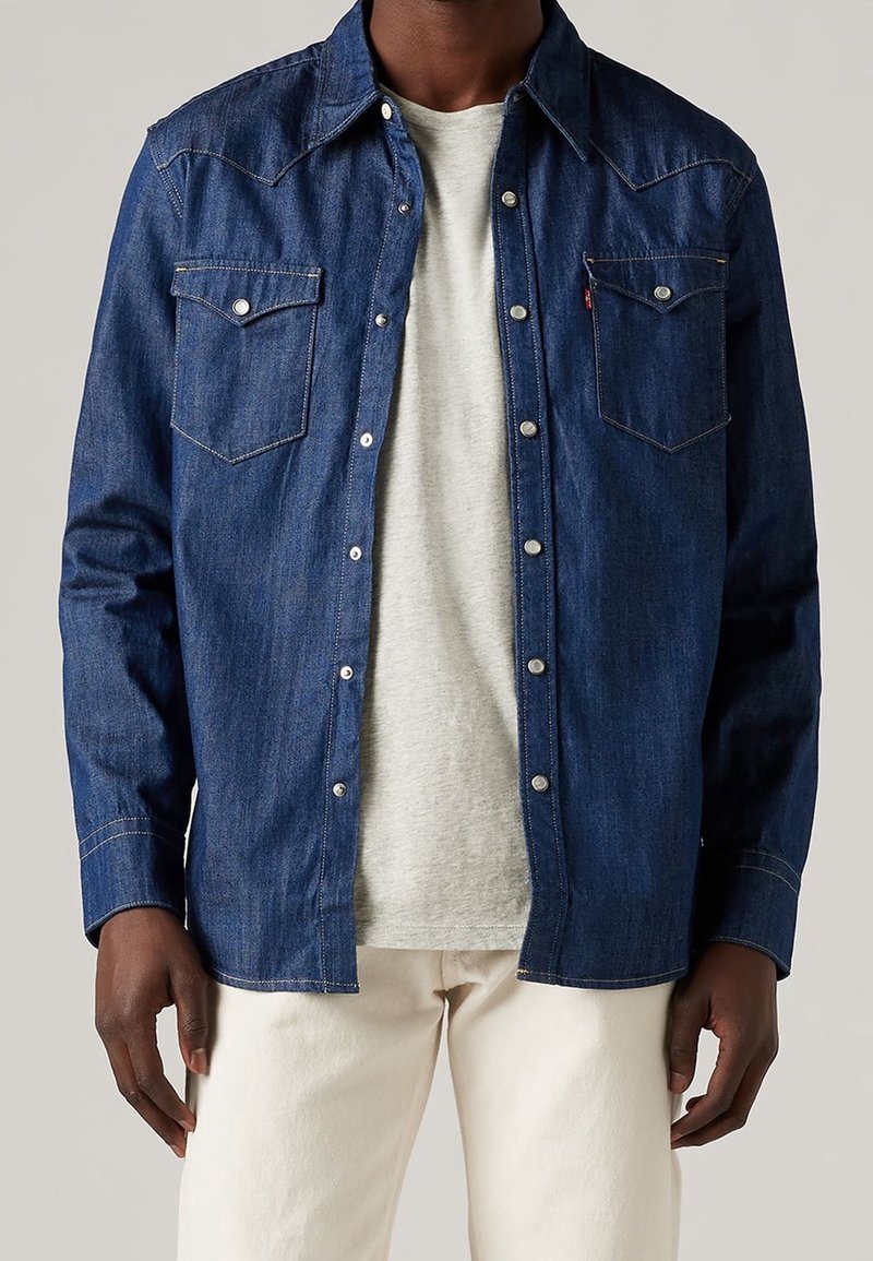 Donkerdenim overhemd met knopen, voorzien van twee borstzakken, zilverkleurige details en contrasterende stiksels, gedragen over een lichtgrijze t-shirt.