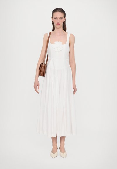 Hvid ærmeløs kjole med blomsterdetalje ved halsudskæringen, A-formet silhuet og en flydende nederdel. Modellen bærer en brun crossbody taske.