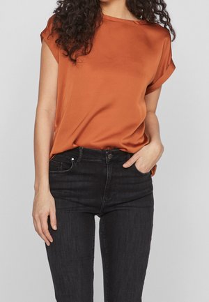 Frau mit lockigem Haar trägt eine rostorangefarbenen Kurzarmbluse, die in eine hoch taillierte schwarze Jeans gesteckt ist, mit einer Hand in der Tasche.