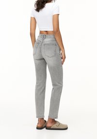 Femme debout portant un jean taille haute gris clair, un haut court à manches courtes blanc, et des chaussures beige à enfiler avec des lanières à boucle.