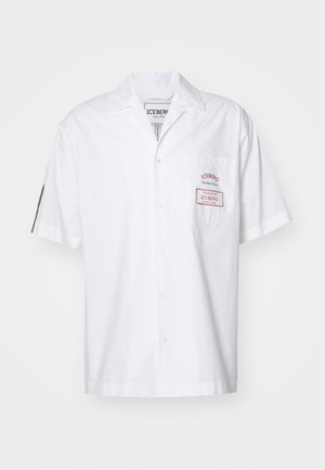 Lühikeste varrukatega valge puuvillane särk, millel on nööpide sulgemine. Omab rinnataskut punase sildiga ja klassikalist kraediagrammi.