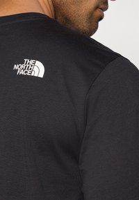 Μαύρο κοντομάνικο T-shirt με ριμπ ύφαση στο λαιμό, που έχει λευκό λογότυπο στο πάνω μέρος της πλάτης. Το ύφασμα φαίνεται λείο με εφαρμοστή σχεδ ίαση.