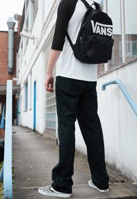Schwarzer Vans-Rucksack, weißes und schwarzes Raglanärmel-Shirt, schwarze Hose und schwarz-weiß karierte Slipper. Im Freien.