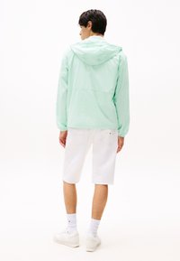 Lichtgroene gewatteerde jas met capuchon en lange mouwen, met elastische manchetten en een relaxte pasvorm, gecombineerd met witte denimshorts en witte sneakers.