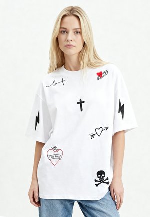 Donna bionda che indossa una maglietta bianca oversize con stampe grafiche nere e rosse che includono un cuore, una croce, un teschio, fulmini e la scritta "Love Hurts."