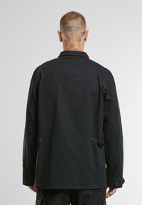 Svart bomullsjacka med spetsig krage, dubbelknappsstängning och texturerade ärmar. Strukturerad, något oversized passform.