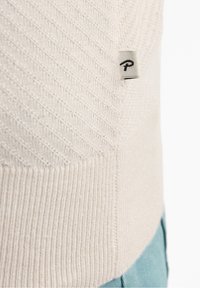 Gros plan sur un pull beige en maille avec un motif diagonal texturé et une petite étiquette en tissu avec un logo, porté sur un pantalon bleu clair.