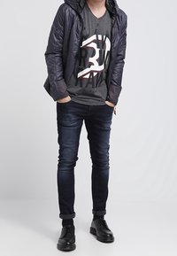 Padded, ciemna kurtka na szarym t-shircie z dekoltem w serek z graficznym wzorem; niebieskie dopasowane jeansy i czarne buty, stojąca na tle w jednolitym kolorze.