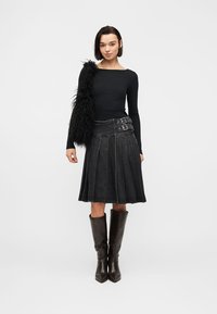 Femme portant un haut noir à manches longues, une jupe plissée noire avec double boucle, un sac d'épaule noir en fourrure et des bottes hautes marron foncé jusqu'aux genoux.