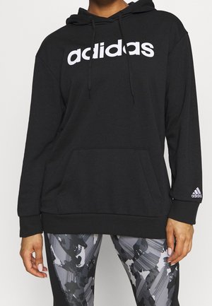 Osoba ubrana w czarną bluzę z kapturem Adidas z białym logo oraz czarne, białe i szare legginsy w wzory na jednolitym białym tle.