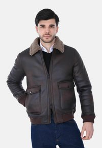 Uomo che indossa una giacca di pelle marrone scuro con colletto in shearling, camicia bianca, maglione nero e jeans blu, in piedi con una mano in tasca.