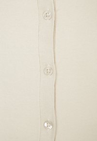 Svetr na knoflíky v krémové barvě, s texturovaným pleteným vzorem a pěti kulatými knoflíky vyrobenými z světlého materiálu.