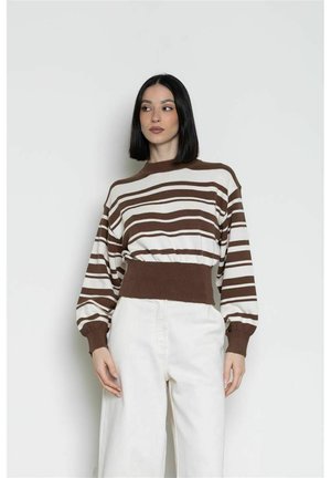 Donna con i capelli neri e lisci che indossa un maglione a righe marrone e bianco e pantaloni bianchi a vita alta, in piedi davanti a uno sfondo semplice.