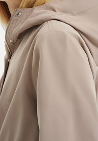 Béžová lehká bunda s kapucí, zapínáním na zip a patentními knoflíky. Hladká látka s jemným leskem, přiléhavý střih a čisté švy.